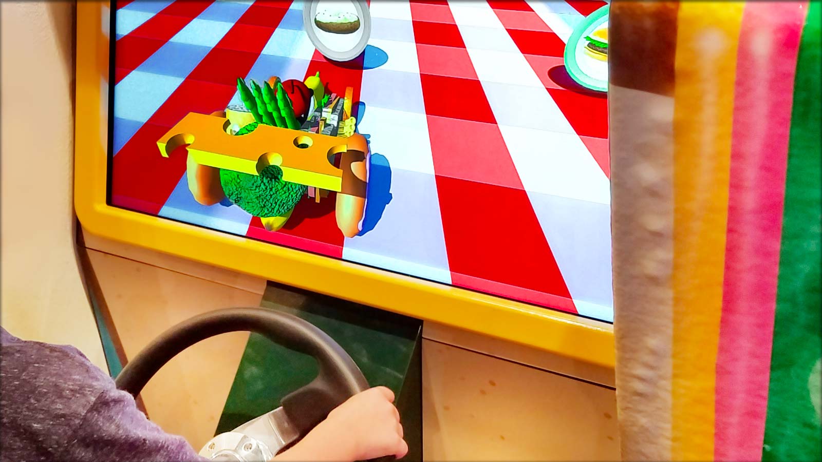 Photo: child using a steering wheel to play driving game in a museum, with a car and obstacles made of food